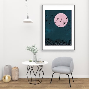 May include: A black framed print of a pink moon with a flock of black birds flying around it against a dark blue background. The moon is in the center of the print and the birds are flying in a circle around it. The print is hanging on a white wall above a white table with a vase of greenery and a chair.