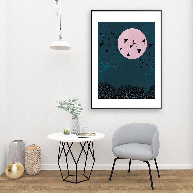 May include: A black framed print of a pink moon with a flock of black birds flying around it against a dark blue background. The moon is in the center of the print and the birds are flying in a circle around it. The print is hanging on a white wall above a white table with a vase of greenery and a chair.