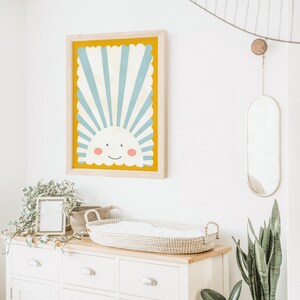 May include: A framed print of a smiling sun with blue and white stripes on a yellow background. The print is hanging on a white wall above a white dresser with a wicker basket on top.