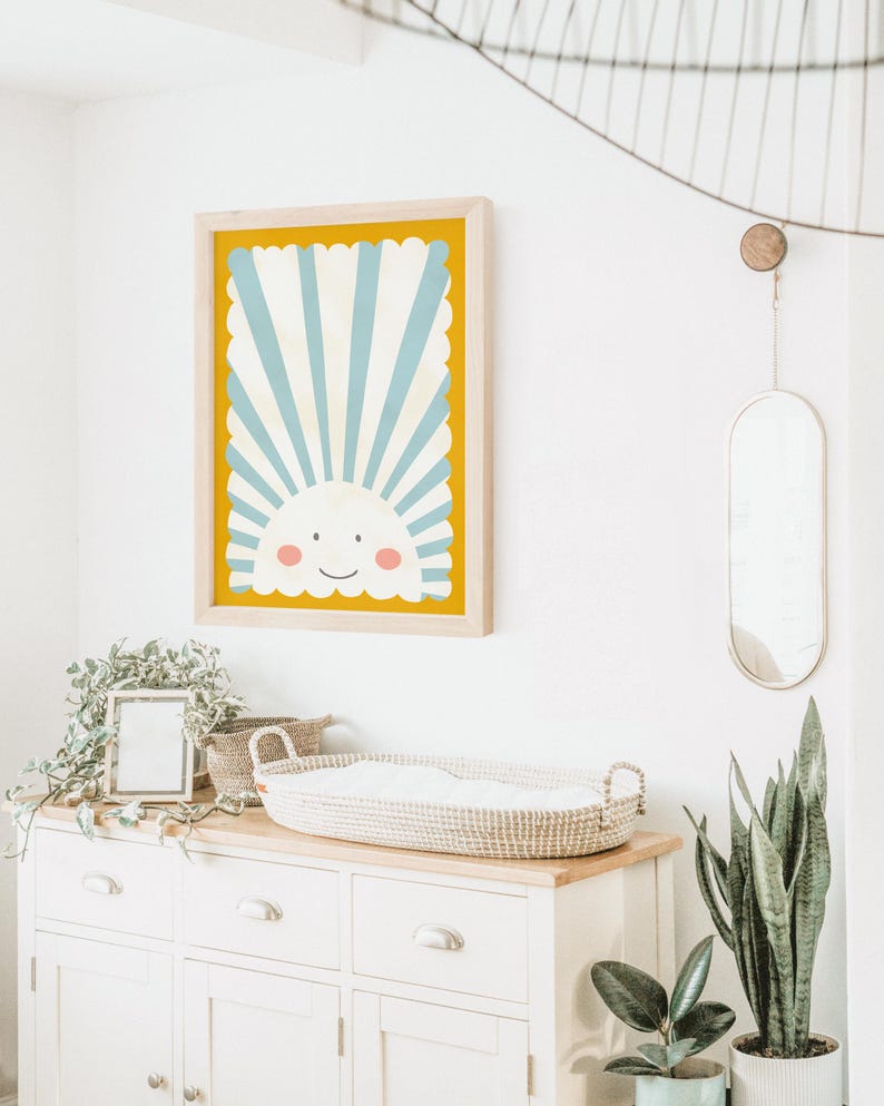 May include: A framed print of a smiling sun with blue and white stripes on a yellow background. The print is hanging on a white wall above a white dresser with a wicker basket on top.