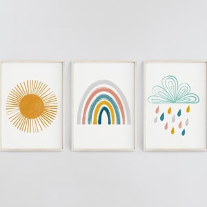 Op de afbeelding: Drie ingelijste prints met een minimalistisch ontwerp. De eerste print toont een gele zon met stralen, de tweede print toont een regenboog met een grijze achtergrond en de derde print toont een blauwe wolk met regendruppels.