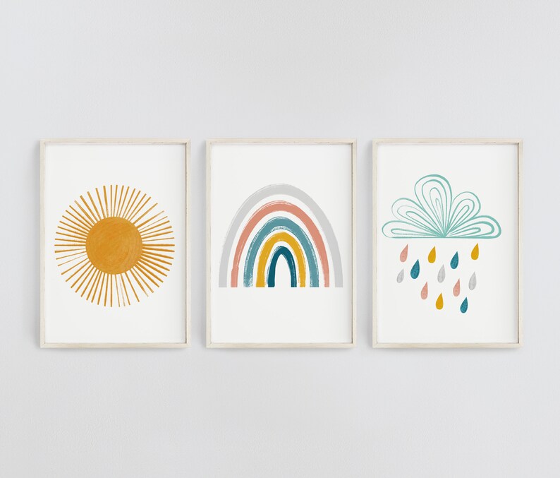 Op de afbeelding: Drie ingelijste prints met een minimalistisch ontwerp. De eerste print toont een gele zon met stralen, de tweede print toont een regenboog met een grijze achtergrond en de derde print toont een blauwe wolk met regendruppels.