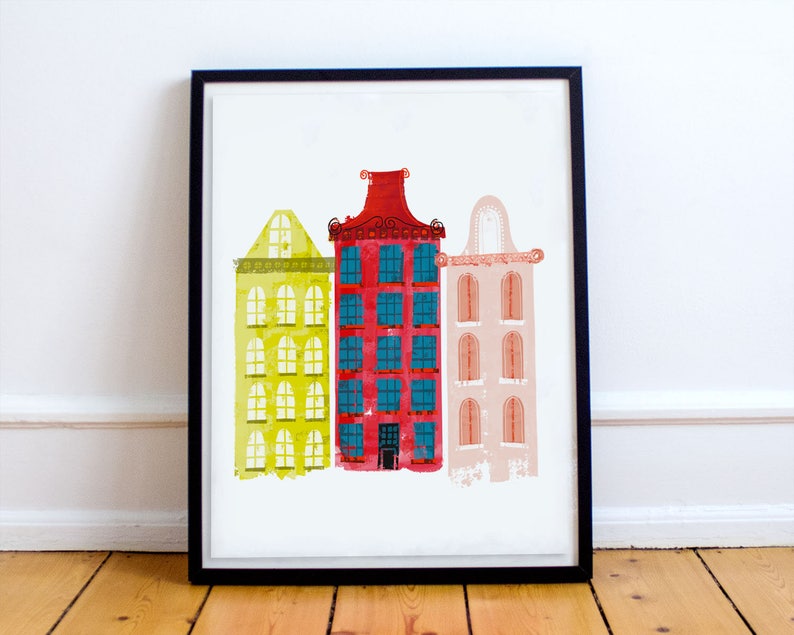 May include: A framed print of three colorful buildings with different architectural styles. The buildings are painted in shades of yellow, red, and pink, with blue windows. The print is a modern and abstract interpretation of a city scene.