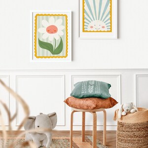 May include: Two framed illustrations, one of a smiling flower and the other of a smiling sun, hang on a white wall. A wooden stool with a green and brown pillow sits in front of a woven basket with a stuffed animal inside.
