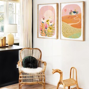May include: Two framed illustrations on a white wall. The illustration on the left features a pink background with a yellow moon and white flowers. The illustration on the right features a pink sky with a yellow sun and a small house on a hill.