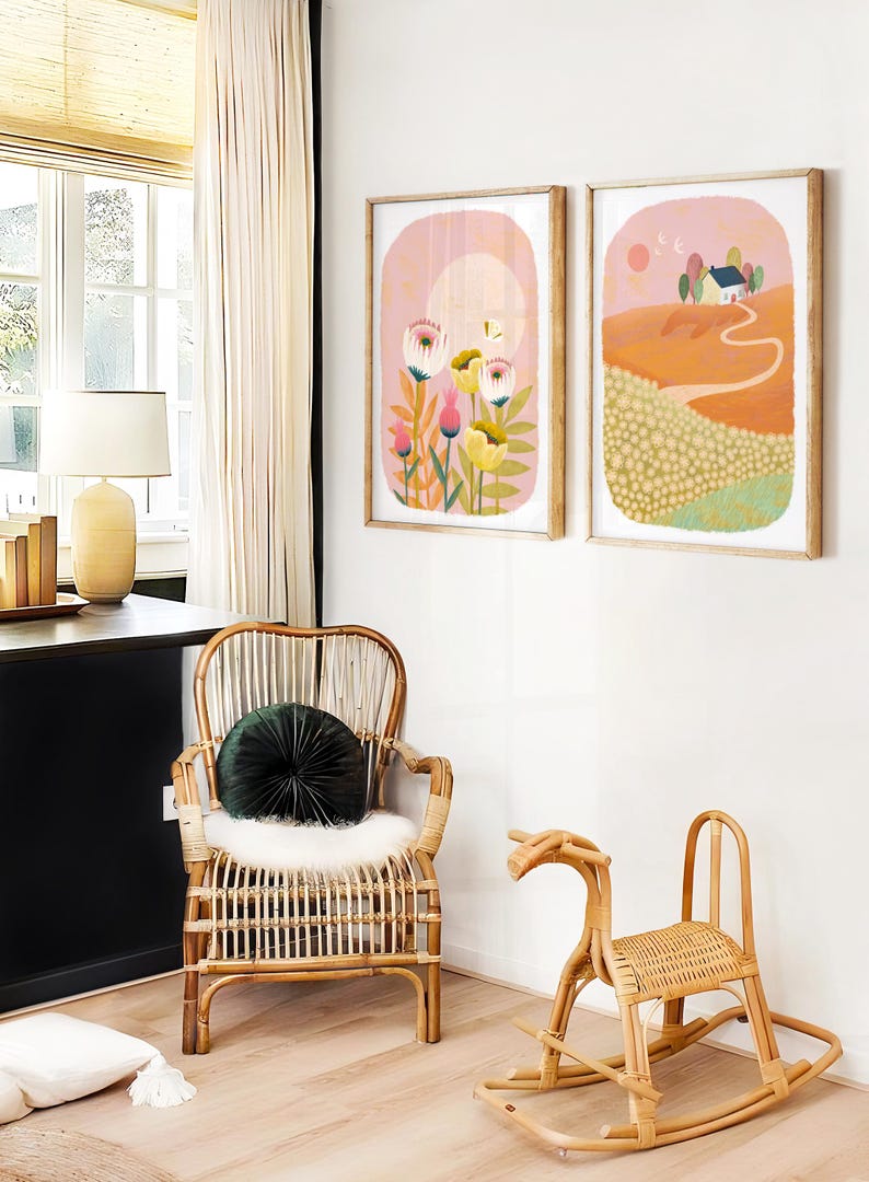May include: Two framed illustrations on a white wall. The illustration on the left features a pink background with a yellow moon and white flowers. The illustration on the right features a pink sky with a yellow sun and a small house on a hill.