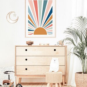 Pu&ograve; includere: Una stampa incorniciata di un design a raggiera colorato con un sole giallo al centro. La stampa &egrave; appesa a una parete bianca sopra una cassettiera in legno con tre cassetti. Un orsacchiotto bianco di peluche &egrave; seduto su uno sgabello intrecciato di fronte alla cassettiera. Una palma in vaso si trova sullo sfondo.