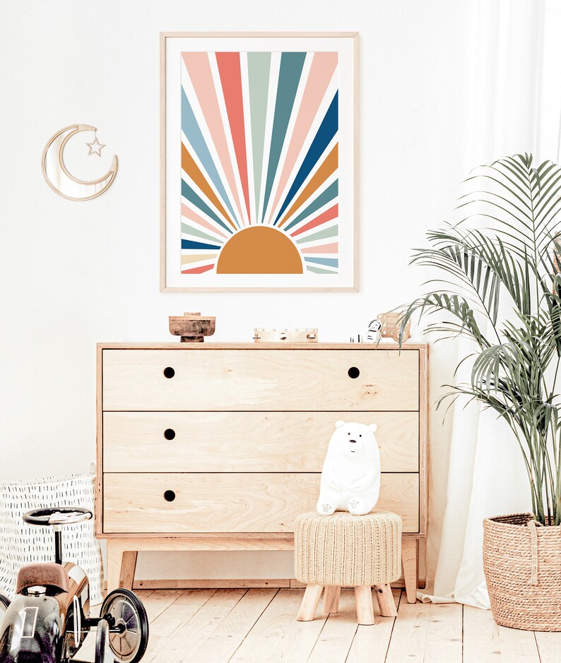 Pu&ograve; includere: Una stampa incorniciata di un design a raggiera colorato con un sole giallo al centro. La stampa &egrave; appesa a una parete bianca sopra una cassettiera in legno con tre cassetti. Un orsacchiotto bianco di peluche &egrave; seduto su uno sgabello intrecciato di fronte alla cassettiera. Una palma in vaso si trova sullo sfondo.