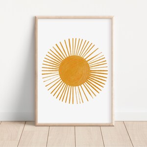 Op de afbeelding: Een ingelijste print van een gestileerde zon in geel-oranje. De zon heeft een cirkel in het midden met stralen die naar buiten uitstralen.