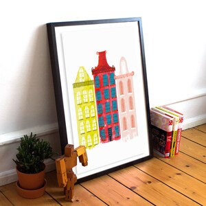 May include: A framed print of a watercolor illustration of three colorful buildings. The buildings are yellow, red, and pink with blue windows. The print is on a white background.