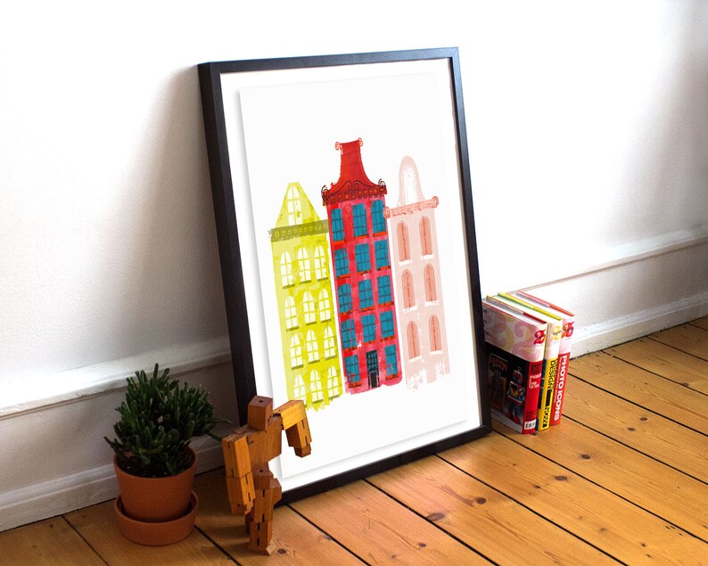 May include: A framed print of a watercolor illustration of three colorful buildings. The buildings are yellow, red, and pink with blue windows. The print is on a white background.