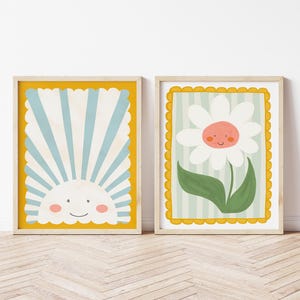 May include: Two framed prints with a yellow border. The print on the left features a blue and white striped sun with a smiling face. The print on the right features a white daisy with a smiling face and green leaves on a light green and white striped background.