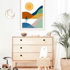 May include: A colorful abstract print of a landscape with a sun, hills, and a sailboat on a body of water. The print is framed in a light wood frame and hanging on a white wall.
