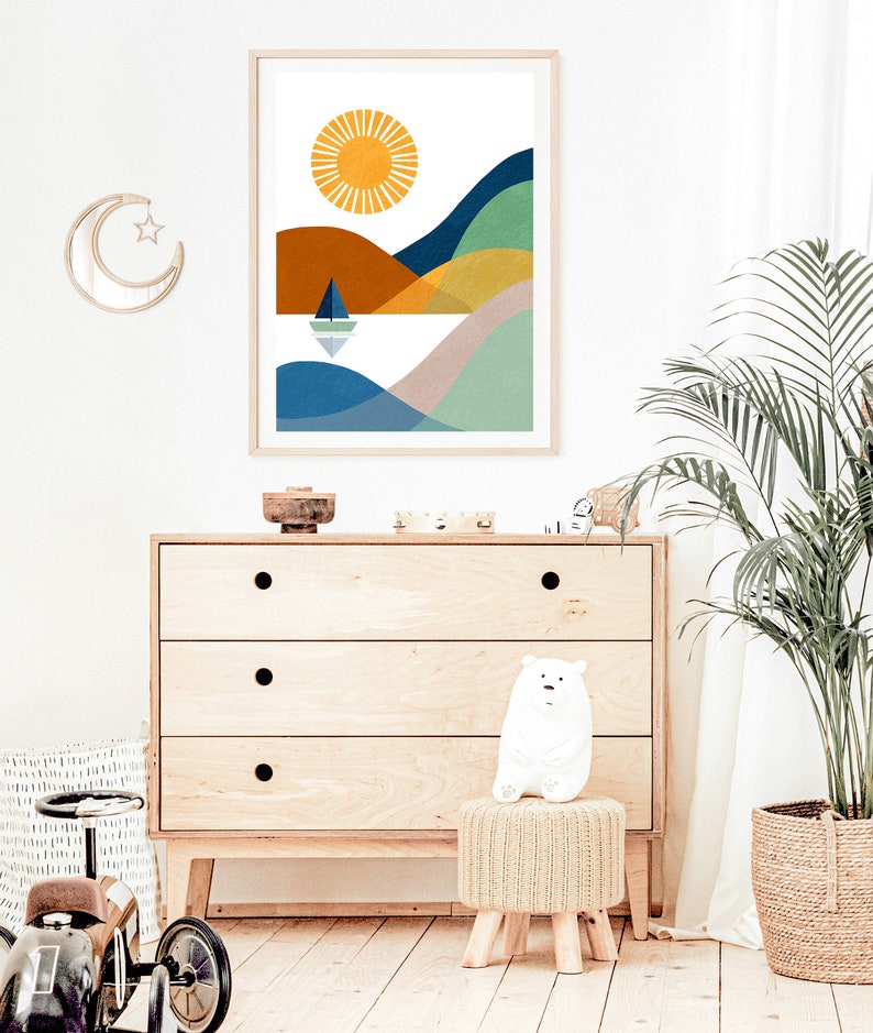 May include: A colorful abstract print of a landscape with a sun, hills, and a sailboat on a body of water. The print is framed in a light wood frame and hanging on a white wall.