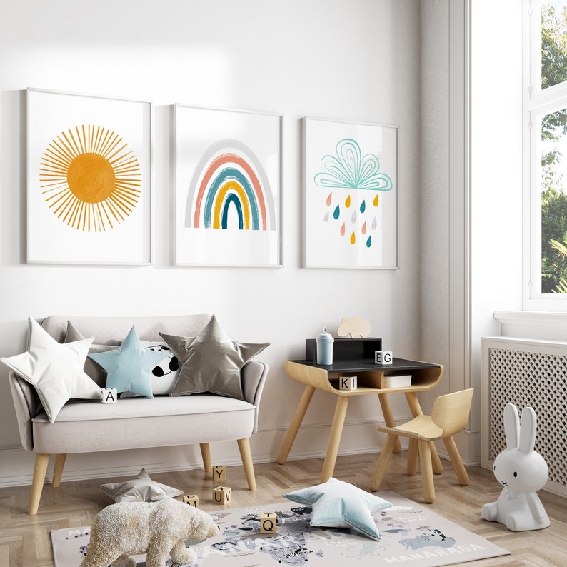 Op de afbeelding: Een lichtgrijze kinderbank met vier kussens, twee grijs, een blauw en een wit met een zwart-wit pandabeerontwerp. De bank staat in een kamer met een houten bureau en stoel, een wit konijnenbeeldje en drie ingelijste prints aan de muur. De prints tonen een gele zon, een regenboog en een blauwe wolk met regendruppels.