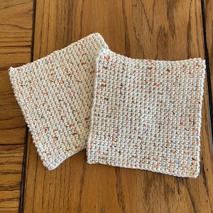 May include: Two square coasters crafted from off-white yarn with a speckled texture. The yarn features small, evenly distributed flecks of orange, blue, and brown throughout the knit pattern, creating a subtle multi-colored appearance.