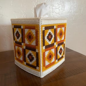May include: A square tissue box cover with a geometric pattern in shades of brown, yellow, and white. The cover is made of a woven material and has a white border. White tissues are visible at the top.