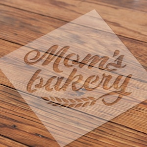May include: A clear, square stencil with the words "Mom's bakery" in a cursive font, with a wheat sprig design below. The stencil is placed on a wooden surface with a warm, brown tone.
