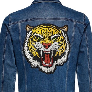 May include: A blue denim jacket with a large embroidered tiger head patch on the back. The tiger has yellow and black stripes, a white face, and a red open mouth.