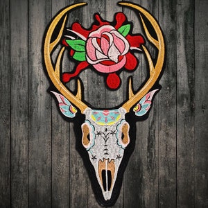 May include: Embroidered patch featuring a deer skull with ornate details, including a pink rose and gold antlers. The skull is white with colorful accents, and the rose has red and green elements. The patch is outlined in black.