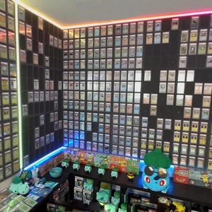 Puede incluir: Una habitación llena de cartas coleccionables enmarcadas, dispuestas en un patrón de cuadrícula en las paredes. Figuras y merchandising de Pokémon se exhiben en estantes debajo. Iluminación LED colorida.