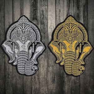 Könnte beinhalten: Zwei gestickte Aufnäher mit der hinduistischen Gottheit Ganesha, einer in Silber und einer in Gold, vor einem dunklen Holz Hintergrund.