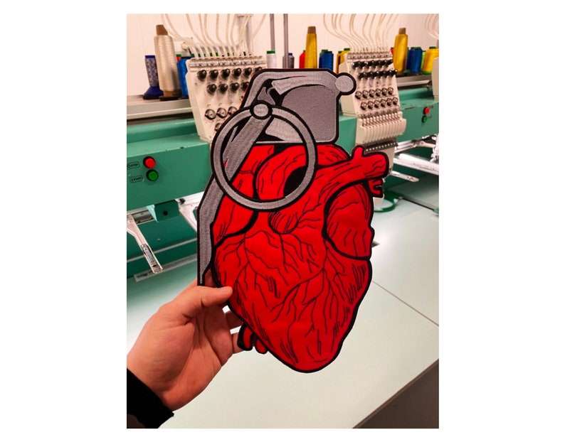 May include: A red embroidered heart with a gray pin attached to it. The heart is being held in front of a sewing machine.