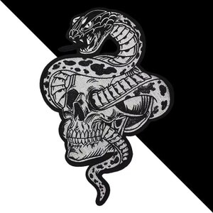 Pode incluir: Um remendo bordado em preto e branco que representa uma caveira com uma cobra enrolada ao redor. A cabeça da cobra está voltada para a frente com a boca aberta. Os olhos da caveira estão fechados. O remendo está sobre um fundo preto e branco.