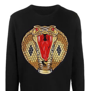 May include: Black sweatshirt with a large embroidered cobra snake design. The snake is gold with red eyes and a red tongue.