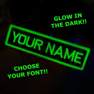 Può includere: Una toppa nera con un contorno verde che brilla al buio. La toppa dice "YOUR NAME" e ha il testo "CHOOSE YOUR FONT!" sotto.