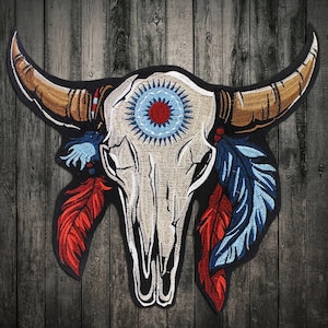 Op de afbeelding: Een geborduurde stieren schedel in wit met rode en blauwe veren. De schedel heeft een blauwe en witte cirkel met een rood midden in het midden van het voorhoofd.