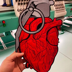 May include: A red embroidered heart shaped like a hand grenade with a silver pin and ring. The heart is detailed with black stitching and is being held by a hand.