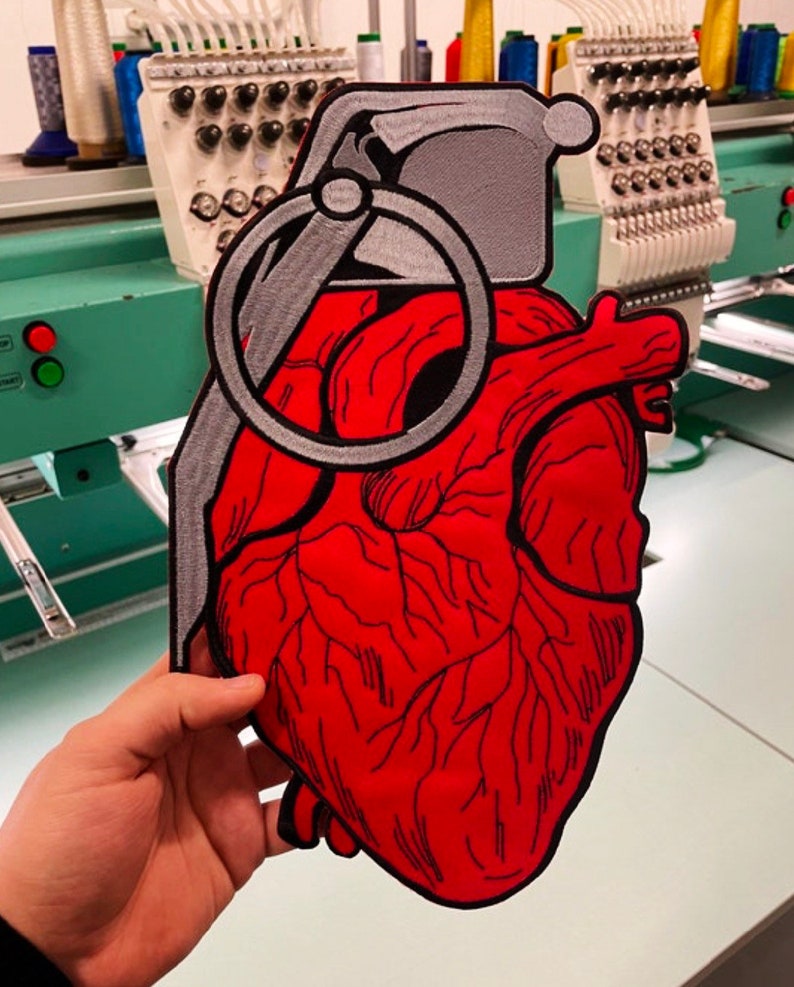 May include: A red embroidered heart shaped like a hand grenade with a silver pin and ring. The heart is detailed with black stitching and is being held by a hand.