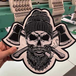 Puede incluir: Parche bordado en blanco y negro de una calavera con un gorro, barba y pipa humeante. Dos hachas cruzadas están detrás de la calavera.