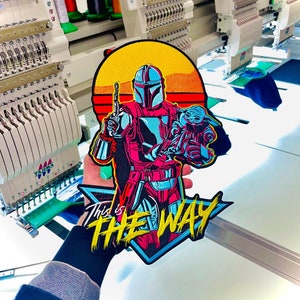 May include: A colorful embroidered patch featuring a character from the Star Wars series, The Mandalorian, holding a small green creature. The patch has a sunset background and the text "This is the Way" in yellow.