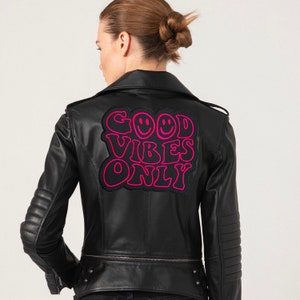 Può includere: Giacca in pelle nera con toppa "GOOD VIBES ONLY" rosa e nera sul retro. La giacca ha un classico stile biker con colletto a revers, zip asimmetrica e dettagli trapuntati su spalle e maniche.
