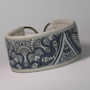 Könnte beinhalten: Ein weißes Lederhundekragen mit einem blauen und weißen gemusterten Stoffeinsatz. Das Halsband hat eine silberne Metallschnalle.