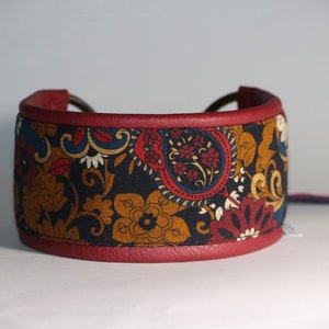 May include: A red leather dog collar with a paisley patterned fabric insert. The fabric is a mix of black, red, blue, and gold colors.