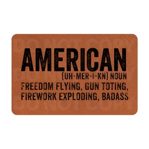 May include: A brown doormat with the word "AMERICAN" in black text. The text below defines "AMERICAN" as a noun and lists characteristics such as "FREEDOM FLYING, GUN TOTING, FIREWORK EXPLODING, BADASS".
