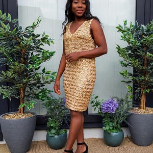 May include: A sleeveless gold sequin dress featuring a V-neckline and a knee-length hem, paired with black block-heeled shoes. The dress is covered in shimmering gold sequins that create a textured, reflective surface across the entire garment.