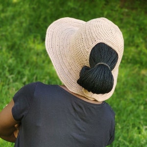 May include: A person wearing a black t-shirt and black pants with a light brown crocheted sun hat.