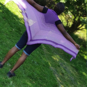 May include: A purple crochet shawl with a gradient of color from light to dark. The shawl is draped over a person's shoulders and is flowing in the wind.