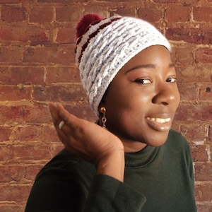 May include: A white and burgundy crochet hat with a pom-pom. The hat has a braided design and is worn by a person. The background is a brick wall.