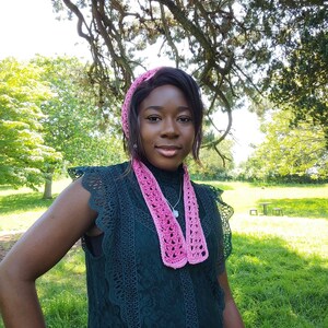 May include: A woman wearing a dark green lace dress with a pink crocheted scarf and headband. She is standing in a grassy area with trees in the background.