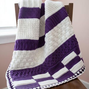 May include: A handmade crochet blanket in white and purple, draped over a wooden chair. The blanket features alternating stripes and squares of white and purple, with textured stitch patterns. The blanket's edge has a decorative border.