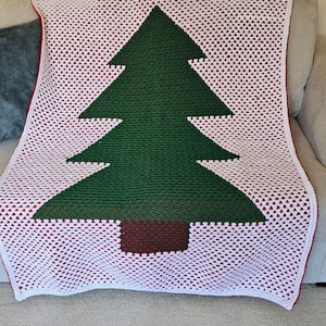 May include: A crocheted blanket featuring a Christmas tree design. The tree is green with a brown trunk, set against a white background with red accents. The blanket is draped over a neutral-colored sofa.
