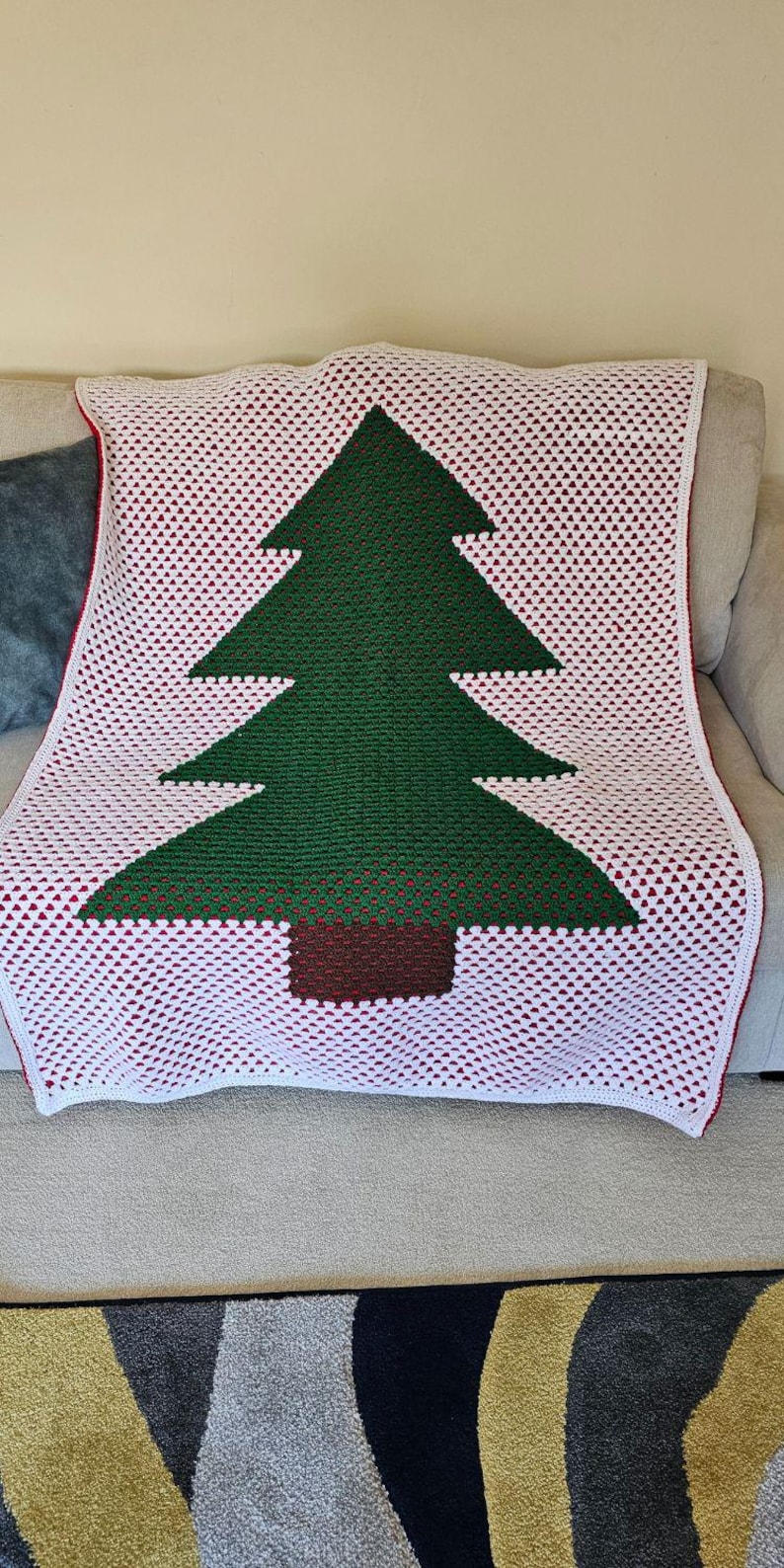 May include: A crocheted blanket featuring a Christmas tree design. The tree is green with a brown trunk, set against a white background with red accents. The blanket is draped over a neutral-colored sofa.