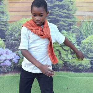 May include: A rust-colored crochet scarf draped around a person's neck. The scarf has a textured pattern and hangs down the front. The person is wearing a white striped t-shirt and black pants. The background features a garden scene.