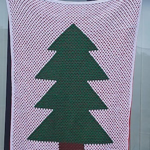 May include: A handmade crochet blanket featuring a green Christmas tree design on a white background with red accents. The blanket is held up, showcasing the tree's shape and the intricate stitchwork. A cozy, festive home decor item.