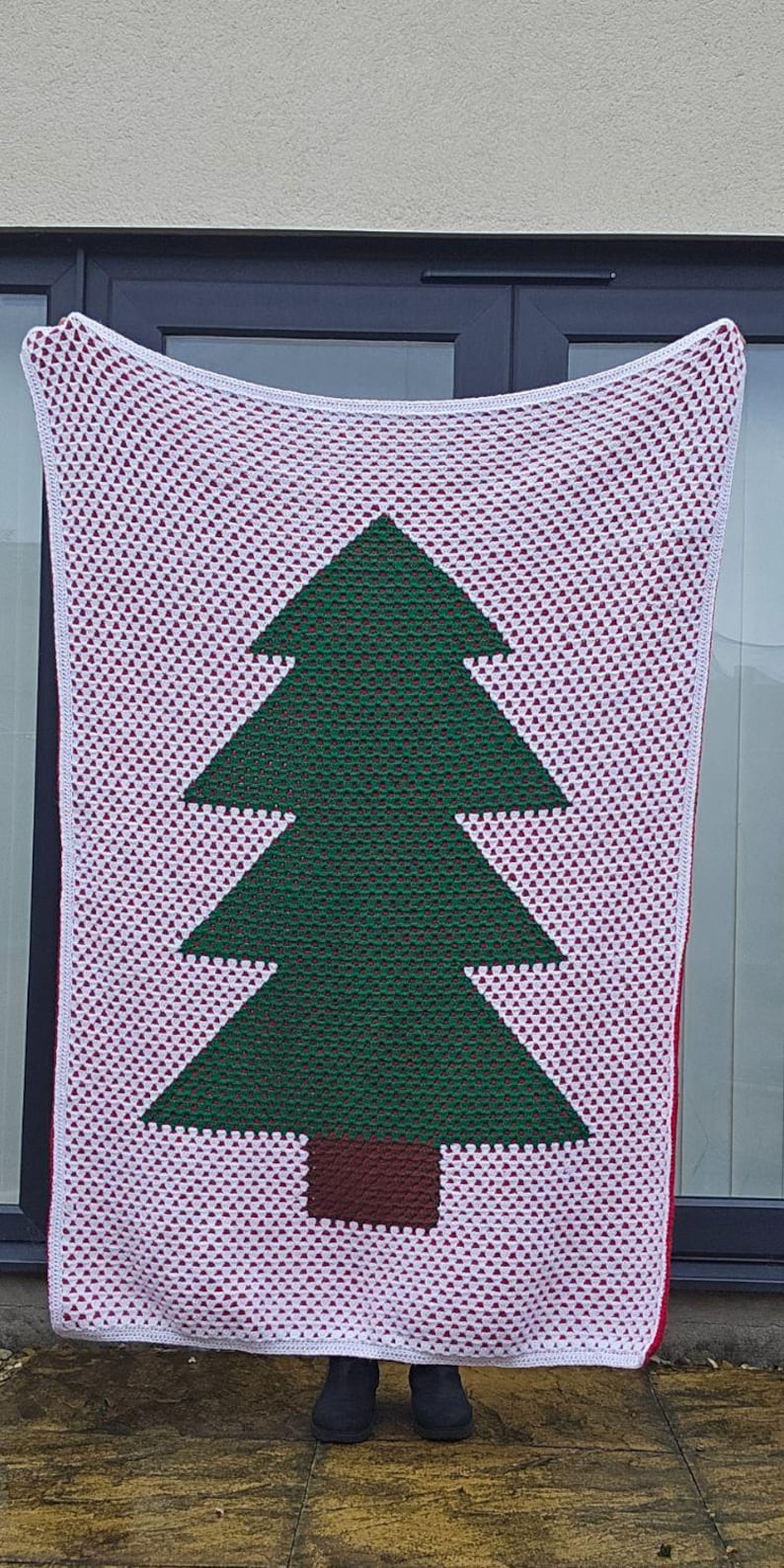 May include: A handmade crochet blanket featuring a green Christmas tree design on a white background with red accents. The blanket is held up, showcasing the tree's shape and the intricate stitchwork. A cozy, festive home decor item.